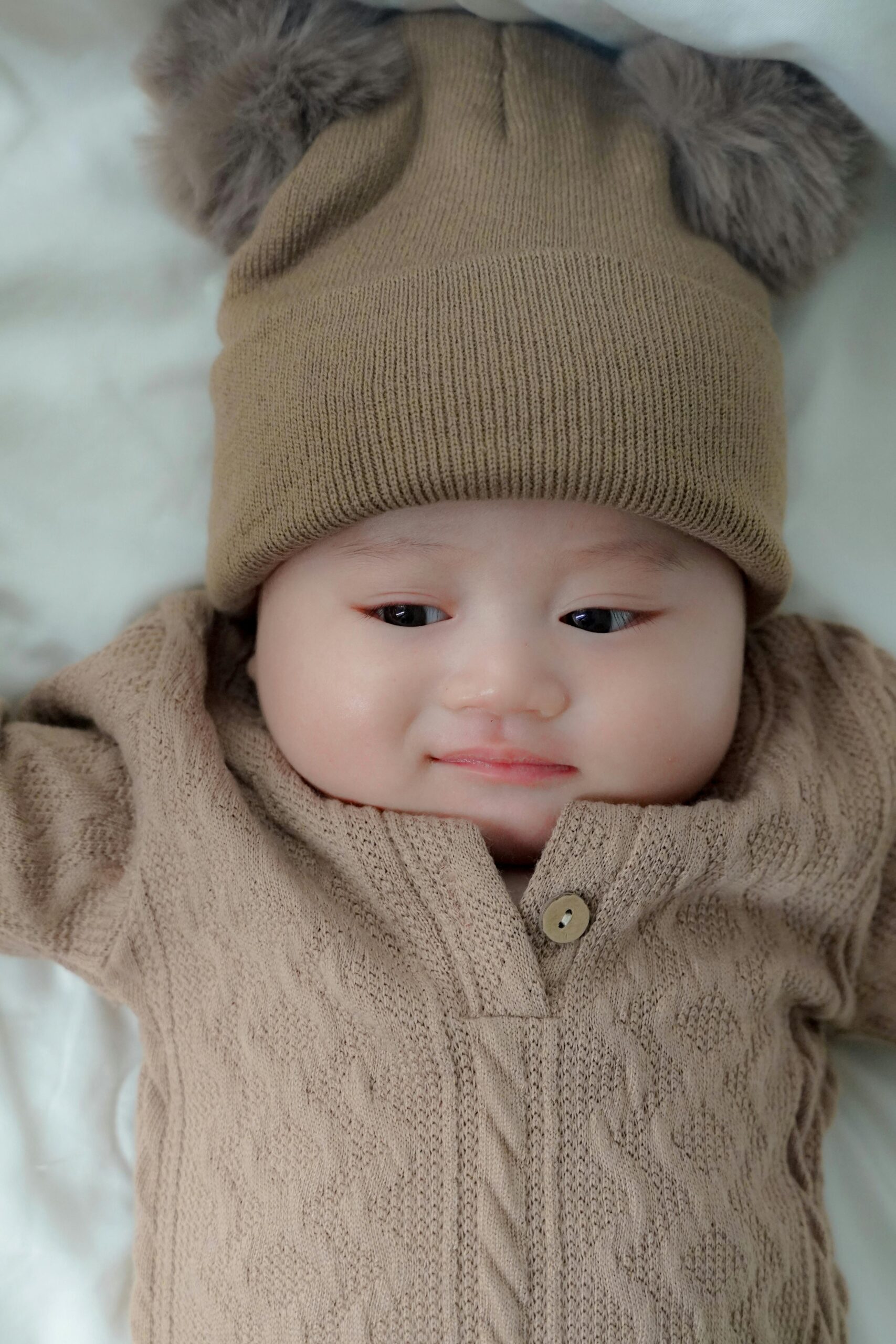 Smiling baby wearing a cozy knit outfit with bear ear hat, lying on a soft white blanket.